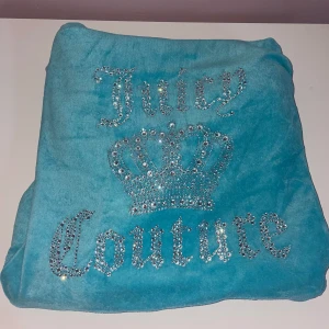 Turkos Juicy Couture zip up - Säljer en turkos zip up från Juicy Couture med huva och glittrande strasstenar på ryggen som bildar text och krona. Jackan har fickor fram och dragkedja. Perfekt för dig som gillar statement-plagg och mjuka material.