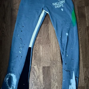 Blå mjukisbyxor från Gallery Dept. - Säljer ett par blå mjukisbyxor från Gallery Dept. med vitt tryck på framsidan och färgstänk i grönt, vitt och svart. Byxorna har dragsko i midjan och en avslappnad passform. Perfekta för en chill och kreativ stil.