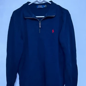 Mörkblå långärmad tröja från Polo Ralph Lauren - Säljer en klassisk mörkblå långärmad tröja från Polo Ralph Lauren med halv dragkedja och röd broderad logga på bröstet. Perfekt för en stilren och avslappnad look. Tröjan har en normal passform och är tillverkad i mjukt material.