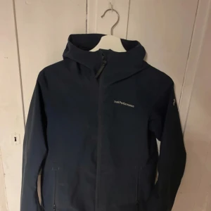 Mörkblå vindjacka från Peak Performance - Säljer en mörkblå vindjacka från Peak Performance med huva och dragkedja framtill. Jackan har två fickor med dragkedja och diskret logga på bröstet. Perfekt för blåsiga dagar och enkel att matcha med olika outfits.