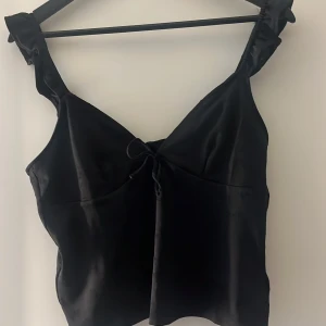 Svart linnetopp med knyt - Snygg svart linnetopp med knyt framtill och breda axelband. Linnet har ett litet mönster vilket är svårt att se på bild och har knytningar i ryggen. Skriv för fler bilder!