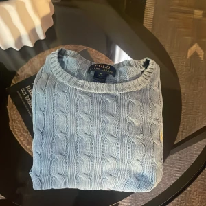 Ljusblå kabelstickad tröja från Polo Ralph Lauren - Säljer en ljusblå kabelstickad tröja från Polo Ralph Lauren. Tröjan har rund halsringning och klassisk kabelstickad design med en liten gul broderad logga på bröstet. Perfekt för en stilren och tidlös look.