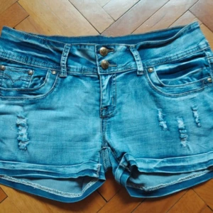 Ljusblå jeansshorts från M-cherry - Säljer ett par ljusblå jeansshorts från M-cherry med slitna detaljer och dubbla knappar framtill. Shortsen har klassiska fickor både fram och bak samt uppvikta benslut. Perfekta för varma dagar!