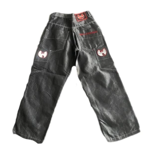 Wu-Tang cargojeans med broderi - Grymma svarta cargojeans med bred passform från Wu-Tang. Byxorna har röda broderade Wu-Tang-loggor på bakfickor och etikett i midjan, samt extra fickor på sidorna. Perfekt för dig som gillar streetwear och vill sticka ut.