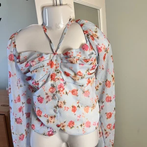 Blommig offshoulder blus - Säljer en söt blommig offshoulder blus i ljusblått med rosa och orange blommor. Blusen har långa ärmar och en knytning framtill. Perfekt för vår och sommar!