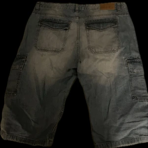 Blå jeansshorts med cargofickor - Säljer ett par blå jeansshorts med cargofickor på sidorna och bakfickor med lock. Klassisk denimlook med avslappnad passform, perfekta för sommaren. Passar dig som gillar en cool stil.