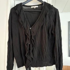 Svart kofta med volang från Flash Woman - Säljer en svart kofta från Flash Woman i storlek XL. Koftan har långa ärmar, v-ringning och dekorativa volangdetaljer längs knäppningen framtill. Tunt och lätt material, perfekt att slänga över en topp.