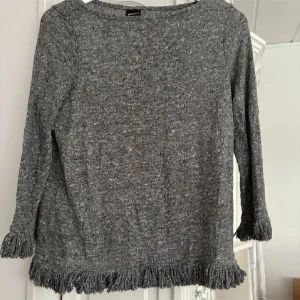 Grå stickad tröja med fransar från Gina Tricot - Säljer en grå stickad tröja från Gina Tricot med långa ärmar och snygga fransdetaljer vid ärmslut och nederkant. Perfekt för dig som vill ha en mysig och trendig look.