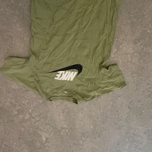 Olivgrön t-shirt från Nike - Säljer en olivgrön t-shirt från Nike med klassisk logga i vitt och svart framtill. T-shirten har rund halsringning och är kortärmad. Perfekt för en sportig och avslappnad stil.    Fåtal gånger använd. Kom gärna med prisförslag om priset inte passar😊