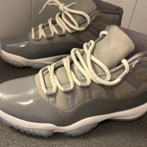 Gråa Air Jordan 11 sneakers - Säljer ett par gråa Air Jordan 11 sneakers med vit sula och vita snören. Skorna har en blandning av blankt och matt material, samt den klassiska Jumpman-loggan på sidan. Perfekta för dig som gillar stilrena och sportiga sneakers.