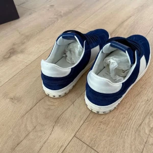 Blå och vita sneakers från Valentino - Säljer ett par snygga blå sneakers från Valentino med vita detaljer och silverfärgade nitar på hälen. Skorna har snörning och är tillverkade i mocka med en vit sula. Perfekta för dig som vill ha en stilren och exklusiv look.