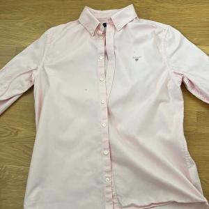 Ljusrosa skjorta från Gant - Säljer en klassisk ljusrosa skjorta från Gant med button-down krage och broderad logga på bröstet. Skjortan har långa ärmar och knäppning framtill. Perfekt för en stilren och avslappnad look.