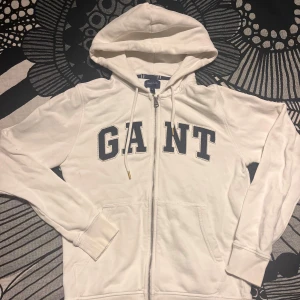 Vit hoodie med dragkedja från GANT - Vit hoodie från GANT med dragkedja framtill och stora broderade bokstäver på bröstet. Tröjan har huva med snörning och två fickor framtill. Perfekt för en avslappnad stil.