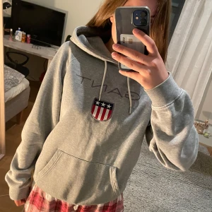 Grå hoodie från Gant - Grå hoodie från Gant med klassisk broderad sköldlogga och text på bröstet. Tröjan har huva med snörning, magficka och långa ärmar. Säljer på grund av att den inte kommer till användning💖