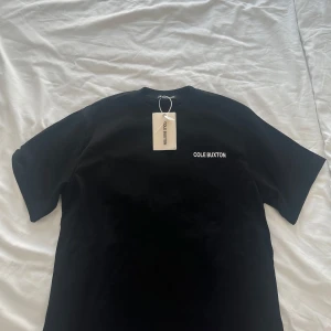 Cule Buxton t-shirt - Svart t-shirt från Cole Buxton med vit logga tryckt på bröstet och stor text på ryggen. Klassisk rund hals och korta ärmar. Tillverkad i mjuk bomull som känns skön mot huden. Perfekt för en clean och stilren streetwear-look. Helt ny! 🤩