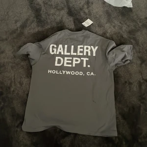 Grå t-shirt från Gallery Dept. - Snygg grå t-shirt från Gallery Dept. med vit logga framtill och stor text på ryggen. Klassisk passform och rund halsringning. Tillverkad i mjuk bomull, perfekt för en clean och avslappnad stil. Helt ny