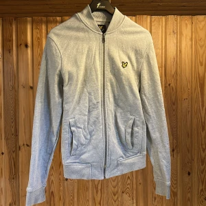 Grå bomberjacka från Lyle & Scott - Snygg ljusgrå bomberjacka från Lyle & Scott med klassisk gul logga på bröstet. Jackan har dragkedja framtill, ribbade muddar och två fickor. Tillverkad i mjuk bomull för en chill och stilren look.