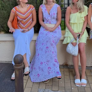 Ljuslila långklänning med volang - Superfin långklänning i ljuslila med blommigt mönster i rosa och lila toner. Klänningen har v-ringning, volangdetaljer vid halsen och är ärmlös. Perfekt för sommarens alla tillfällen och ger en romantisk vibe.