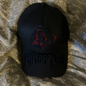 Svart keps från Philipp Plein - Snygg svart keps från Philipp Plein med broderad döskalle och logga framtill samt broderad logga baktill. Justerbar rem med metallspänne. Kepsen har en cool streetstil och är tillverkad i ett glansigt syntetmaterial.