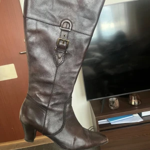 Boots stövletter mörkbruna - Snygga mörkbruna stövlar i skinn med dekorativt spänne och markerade sömmar. Modellen har spetsig tå och stabil klack som ger en dressad look. Perfekta till höst- och vinteroutfits för dig som gillar klassisk stil.
