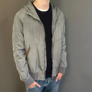 Grå vindjacka från Tom Ford med huva - Snygg grå vindjacka från Tom Ford med huva och dragkedja framtill. Jackan har ribbade muddar, två fickor med knappdetalj och en cool spänndetalj på ryggen. Tillverkad i Italien och fodrad med mesh för extra komfort. Perfekt för dig som gillar stilren och sportig look.