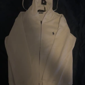 Helt nya Vit zip up hoodie från Polo Ralph Lauren - Tjenare jag säljer en Snygg vit hoodie från Polo Ralph Lauren med dragkedja och klassisk huva. Tröjan har det ikoniska broderade logotypen på bröstet och är i mjuk bomull. Perfekt för en clean och stilren look. Långärmad och med snörning i huvan. DEN ÄR HELT NY OCH ALDRIG ANVÄND!!!