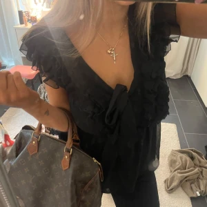 Svart blus med volang och knyt - Supersöt svart blus i luftig polyester med volangdetaljer längs v-ringningen och ärmarna. Djup v-ringning och stor knyt framtill 