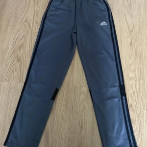 Grå Adidas mjukisbyxor med svarta ränder - Grå mjukisbyxor från Adidas med svarta ränder längs sidorna och breda svarta detaljer vid knäna. Byxorna har elastisk midja och är tillverkade i 100% polyester, perfekta för träning eller chill. Klassisk Adidas-logga framtill.