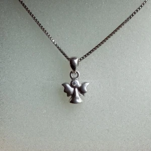 Halsband i äkta silver med ängel - Sött halsband i äkta 925 silver med en liten ängel som hänge. Ängeln har en rund glittrande detalj i mitten och hänger på en tunn, fyrkantig kedja. Perfekt för dig som gillar stilrena och symboliska smycken.