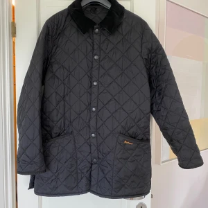 Svart quiltad jacka från Barbour - Quiltad svart jacka från Barbour med klassisk krage i manchester och tryckknappar framtill. Jackan har två stora fickor med broderad Barbour-logga och är helsvart med rutmönster. Perfekt för dig som vill ha en stilren och tidlös look.