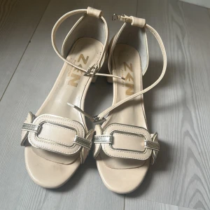 Beige sandaler med blockklack från ZEN - Stilrena beige sandaler från ZEN med öppen tå, blockklack och smala ankelremmar med spänne. Snygga detaljer i silver och mjukt syntetmaterial. Perfekta för dig som gillar en clean och modern look.