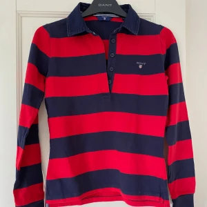 Randig pikétröja från GANT XS - Snygg och cool långärmad pikétröja från GANT i marinblått och rött med breda ränder. Klassisk krage, knappar framtill och broderad logga på bröstet. Tillverkad i mjuk bomull.