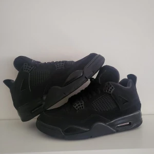 Svarta Nike Air Jordan 4 sneakers - Säljer ett par helsvarta Nike Air Jordan 4 sneakers med klassisk meshpanel, snörning och ikoniska Jumpman-loggan på plösen och sulan. Skorna har en robust sula med synlig Air-enhet och ovandel i mocka och syntet. Perfekta för dig som gillar stilrena och sportiga sneakers.