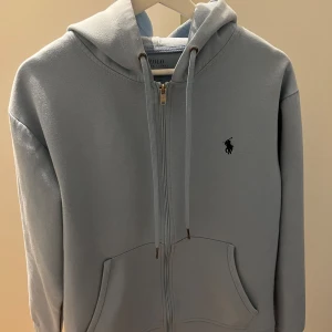 Polo Ralph Lauren - Snygg ljusblå hoodie från Polo Ralph Lauren med dragkedja och klassisk huva. Tröjan har fickor framtill och den ikoniska broderade loggan på bröstet. Perfekt för en chill och stilren look.