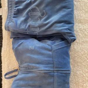 Blå Juicy Couture mjukisbyxor - Säljer ett par blå mjukis set från Juicy Couture i mjuk velour med resår och dragsko i midjan. Byxorna har fickor och dekoreras med glittriga stenar, samma med ryggen på tröjan. De är i väldigt bra skick och är i storlek XS