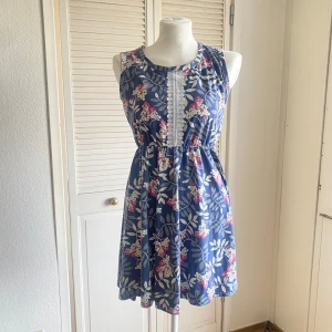 Blommig blå kortklänning med spets - Supersöt blå sommarklänning med blommigt mönster i vitt och rosa. Ärmlös modell med resår i midjan och dekorativ vit spets framtill. Perfekt för varma dagar och har en avslappnad, flowy passform.