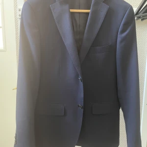 Mörkblå kavaj från Riley slim fit - Snygg mörkblå kavaj från Riley i slim fit-modell. Klassisk design med två knappar framtill, fickor med lock och bröstficka. Perfekt för dig som gillar en stilren och modern look. Fodrad insida för extra komfort.