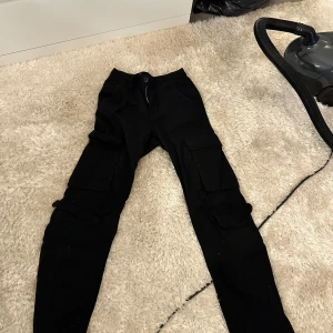 Svarta cargopants med fickor - Svarta cargopants med flera stora fickor på benen och smal passform. Perfekta för en streetwear-look och riktigt bekväma med sin mjuka bomullskänsla. Stängs med knapp och dragkedja framtill.
