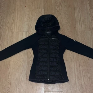 Svart pufferjacka Peak Performance XS - Svart pufferjacka från Peak Performance i storlek XS. Jackan har quiltad front, huva och hel dragkedja. Snygg och sportig look med diskret logga på bröstet och ärmen. Perfekt för kyliga dagar när du vill hålla stilen.