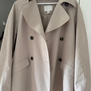Beige trenchcoat från H&M, stl M - Snygg beige trenchcoat från H&M i klassisk modell med dubbelknäppning, stora slag och bälte i midjan. Jackan har långa ärmar och två fickor framtill. Aldrig använt, köpt för 800kr säljer för 400kr. Pris kan diskuteras. 