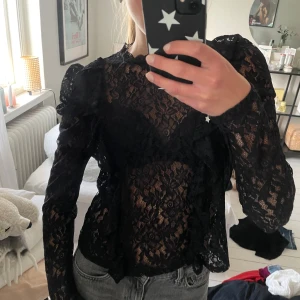 Svart spetsblus med volangdetaljer - Säljer en svart långärmad blus i spets med blommigt mönster och volangdetaljer på axlar och framtill. Den är oanvänd men saknar lappar , skriv om fler bilder eller frågor.