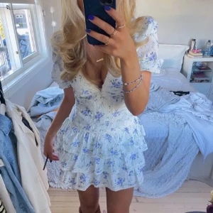 Zara liknande klänning  - Supersöt vit kortklänning med blått blommönster, puffärmar och v-ringning. Klänningen har flera lager volanger och spetsdetaljer längs kanterna. Perfekt för sommarens alla tillfällen och ger en romantisk vibe. Aldrig använd. 