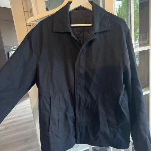 Navy ulljacka från Peak Performance - Snygg navy jacka från Peak Performance i ullblandning. Klassisk rak modell med krage, dolda knappar och två fickor framtill. Fodrad insida för extra komfort. Perfekt för dig som gillar stilren och enkel design. Perfekt för svalare sommarkvällar, vår eller höst. 