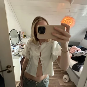 Vit blus med knyt och puffärm - Superfin vit blus med korta puffärmar och två knyt framtill som ger en öppen look. Blusen är croppad och har en luftig känsla, perfekt för varmare dagar. Enkel men ändå unik med sina knyt-detaljer. Det har tyvärr sprättats upp lite vid ryggen🥲men inget som syns synligt 