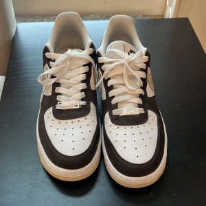 Nike Air Force 1 brun/beige sneakers - Snygga Nike Air Force 1 sneakers i brunt och vitt skinn med beige swoosh och detaljer. Klassisk siluett med perforerad tå, vit sula och snörning. Perfekta för dig som vill ha en clean och tidlös look med streetkänsla.