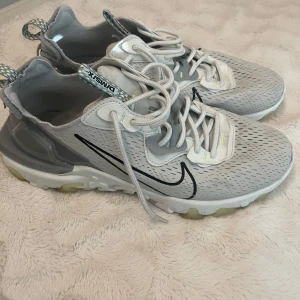 Nike React Vision - Nike React Vision sneakers i ljusgrått med svarta detaljer och genomskinliga partier. Snygg design med mönstrad dragflik bak, vit sula med reakt-teknologi och snörning framtill. Perfekta för dig som gillar streetstyle och modern look.