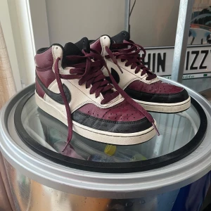 Nike skor vinröd/svart/vit - Nike sportswear Court Vision Low sneakers i vinrött, svart och vitt skinn. Klassisk high-top siluett med svart swoosh, vinröda skosnören och perforerad tå. Tydlig retrostil och ikonisk design som sticker ut. Perfekta för dig som gillar streetwear och sneakers med attityd. Bra skick använd men inga skador eller liknande. Stått i skohyllan ca 2 år så säljer nu