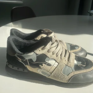 Valentino Rockrunner sneakers camo - Snygga Valentino Rockrunner sneakers med beige mockadetaljer, svart och grått camouflagemönster samt svarta nitar på hälen. Skorna har snörning, platt sula och rund tå. Perfekta för dig som gillar streetstyle och exklusiva märken.