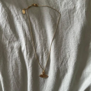 Guldigt halsband Vivienne Westwood - Säljer ett ikoniskt guldigt halsband från Vivienne Westwood med den klassiska orb-berlocken. Kedjan är tunn och elegant, och halsbandet kommer med originalask och tygpåse. Perfekt statement-piece för dig som gillar coola accessoarer.