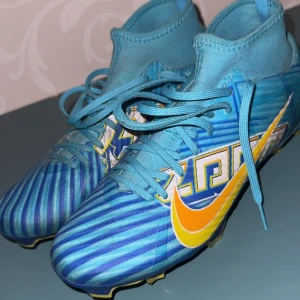 Nike mercurial fotbollsskor - Snygga Nike Mercurial fotbollsskor i blått med randigt mönster och orange-gul swoosh. Skorna har snörning, högre strumpliknande skaft och mönstrad sula med dobbar för gräsplan. Perfekta för dig som vill sticka ut på planen.